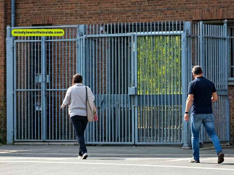 Two people stroll by a metal gate, likely near a prison or secure area, suggesting a visit to an inmate.