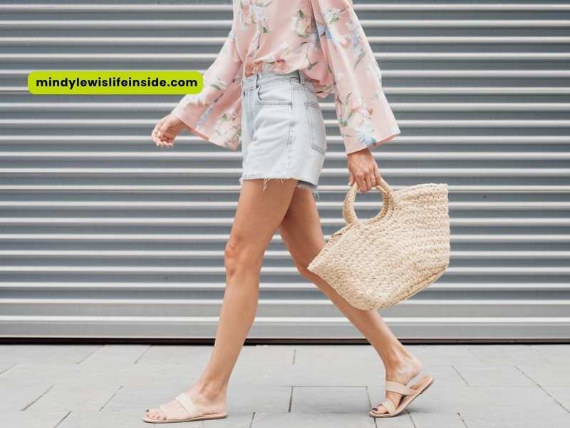 A woman wearing denim shorts and a pink floral top strolls along the street, showcasing a casual summer outfit.