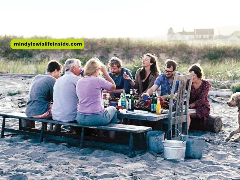 A group of friends enjoying a meal together at a picnic table on the beach, with the ocean in the background.