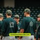 Baseball players in green uniforms engage in conversation, showcasing their camouflage hats during a themed game.