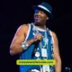 On stage, a man in a blue shirt and hat captivates the audience, reminiscent of Slick Rick's iconic eyepatch look.