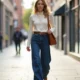 A woman wearing a white sweater and loose jeans walks casually along a street, surrounded by buildings.