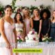 Six smiling women in elegant dresses stand around a floral-decorated table with a cake. Pink balloons and lush greenery create a festive, joyful atmosphere.