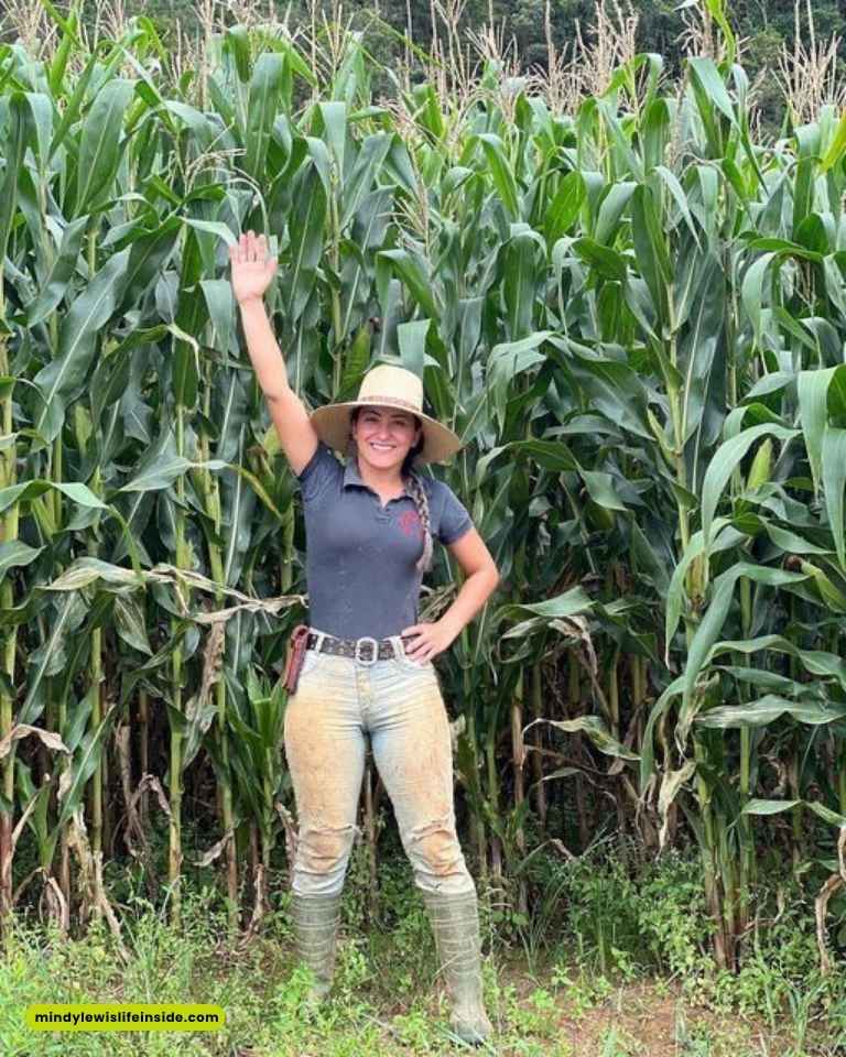A woman wearing a fashionable hat poses in front of a vibrant cornfield, showcasing a blend of style and rural charm.