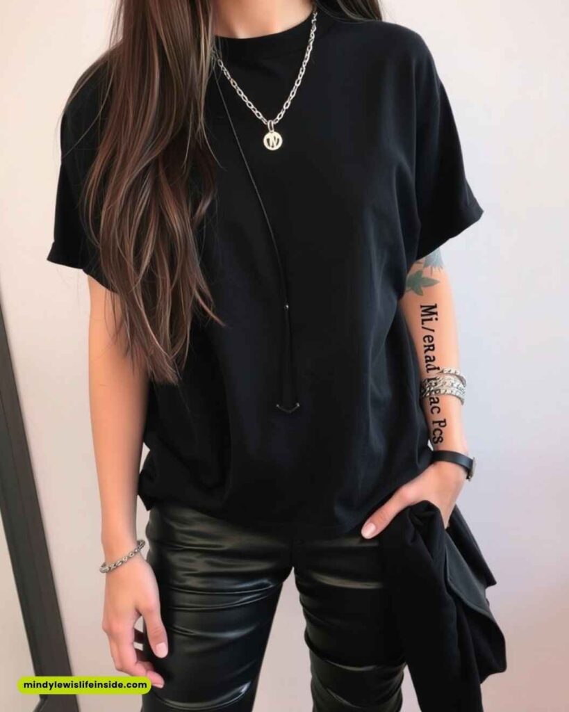 Young woman wearing a black t-shirt and leather pants stands with one hand in her pocket. Accessories include a silver chain necklace and bracelets. Modern and stylish.