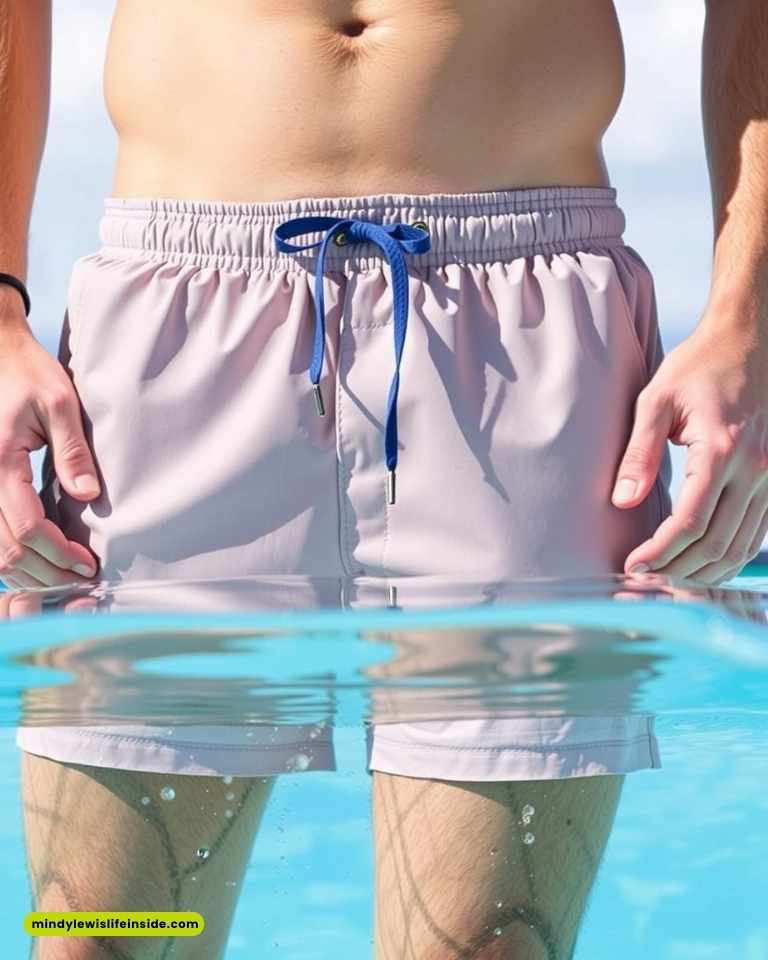 A person stands waist-deep in clear blue water, wearing light gray swim trunks with a blue drawstring. The scene suggests a relaxed, sunny beach day.