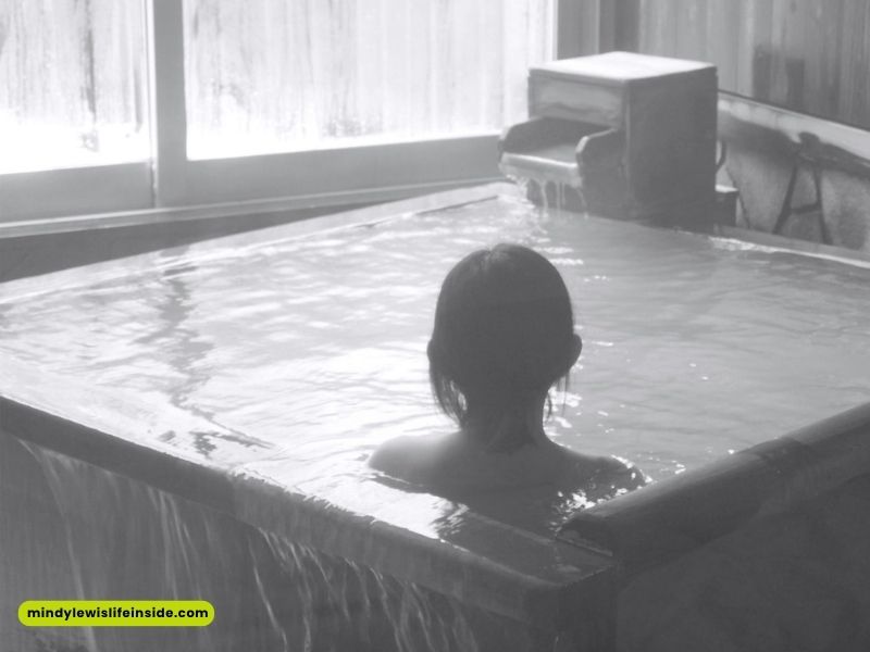 A woman sits in a bathtub with water flowing over the edges, pondering the rules of wearing swimsuits in onsens.
