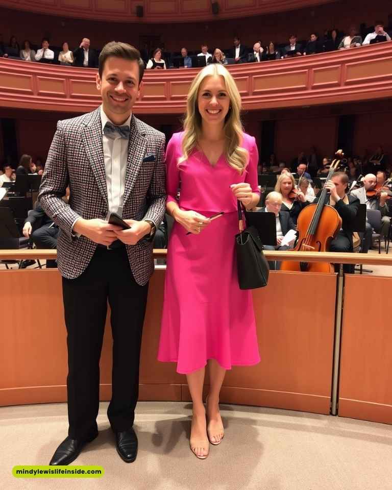 A smiling couple stands in a concert hall; the man wears a checkered blazer and bow tie, and the woman in a bright pink dress, with an orchestra behind them.