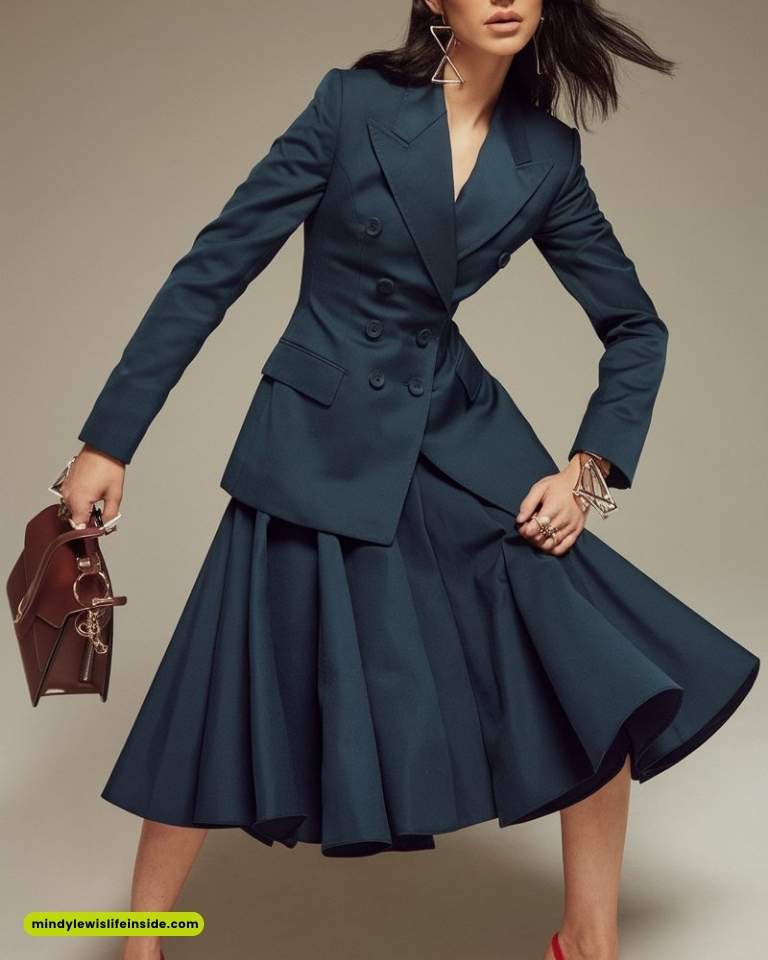A person poses in a stylish dark teal suit with a fitted double-breasted blazer and flared skirt. They hold a brown handbag and wear geometric earrings.