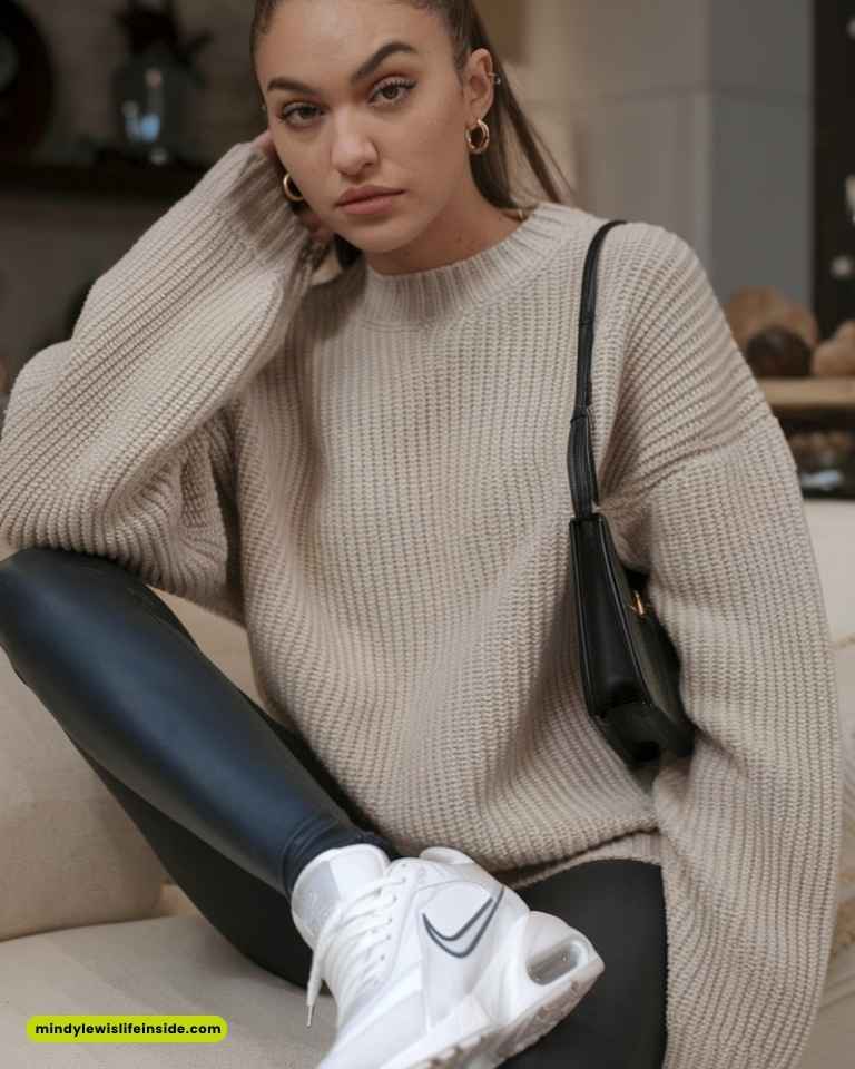 Woman in a cozy beige sweater and black leather leggings sits casually indoors. She wears white sneakers and carries a black shoulder bag, conveying a relaxed, stylish vibe.