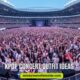 A massive crowd fills a stadium, cheering with glow sticks. The stage is in the distance. Text overlay reads "Kpop Concert Outfit Ideas." The atmosphere is energetic.